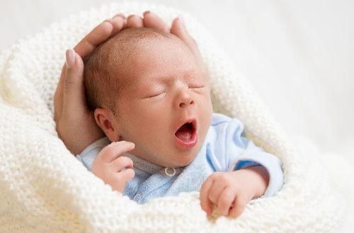 Newborn baby yawning with a hand gently holding its head, lying on a soft white blanket.